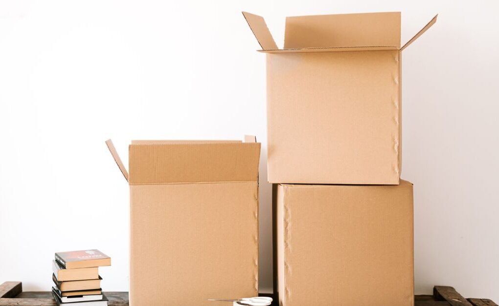 Opened cardboard boxes and stack of books placed on wooden shabby table near tape with scissors against white plain wall